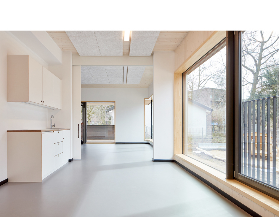 Aloys Kiefer Architekturfotografie: Querformat, sehr helle Innenansicht. Durchblick auf hinteren Räume. Rechts, Durchblick durch ein großes Fenster in den Außenbereich. Links eine Pantryküche mit Holzdekor.