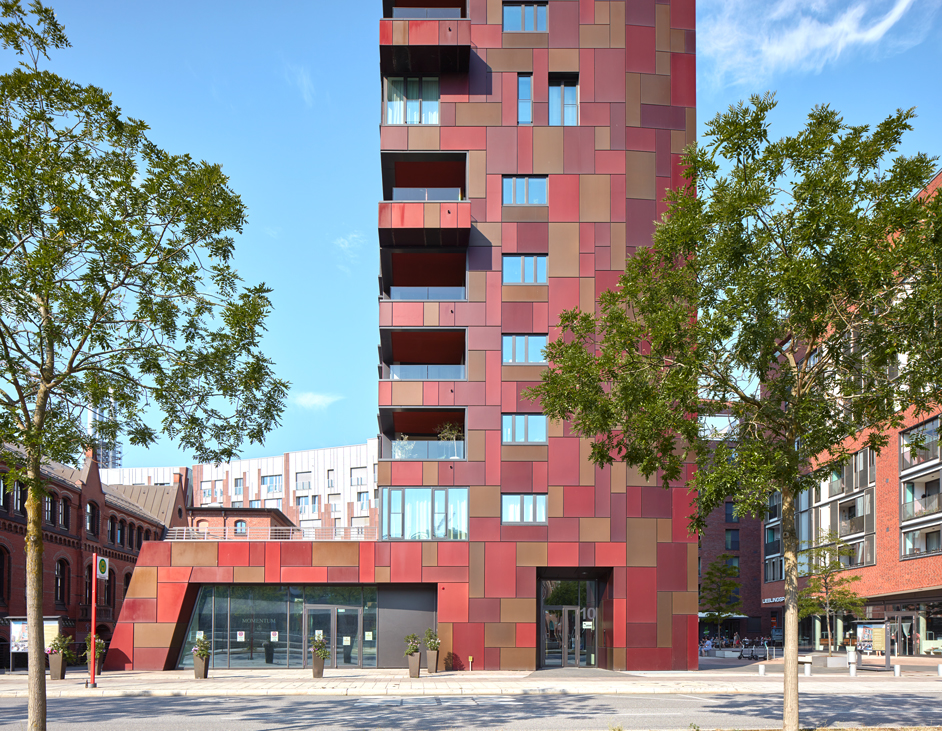 Aloys Kiefer Architekturfotografie: Motiv im Querformat. Im Vordergrund rechts und links Bäume mit Blättern. Im Hintergrund ein rotes Hochhaus mit Balkonen.