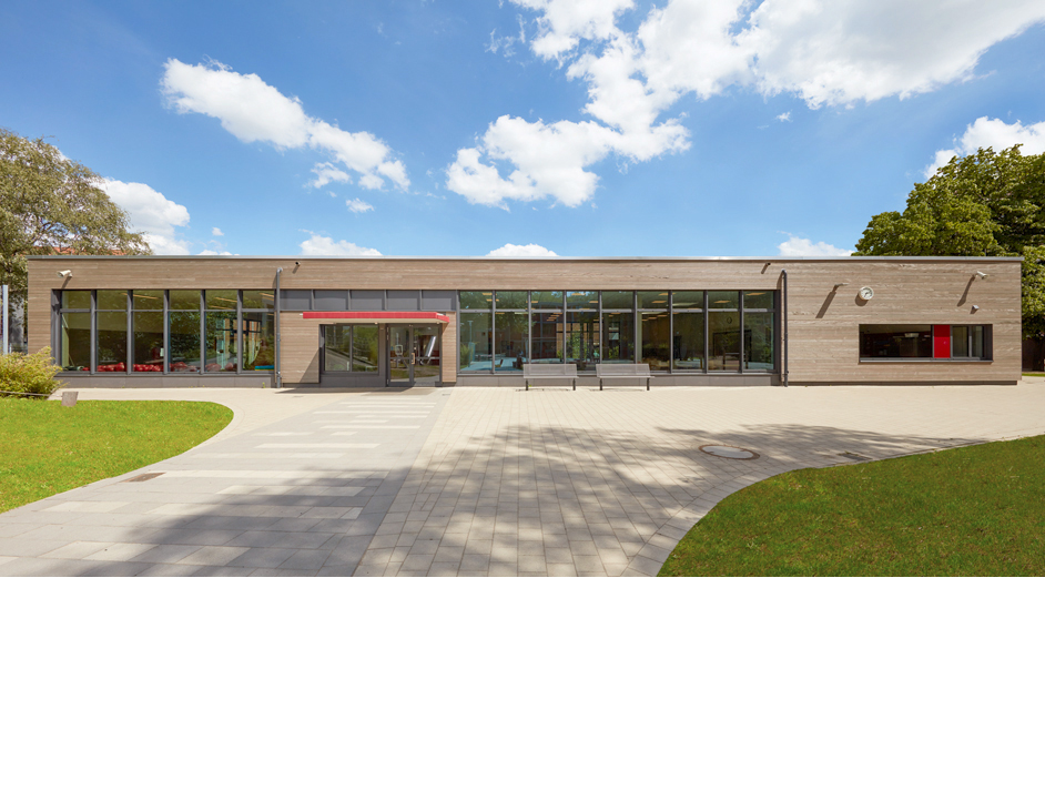 Aloys Kiefer Architekturfotografie: Querformat, im Vordergrund rechts und links Rasen in der Vordergrund-Mitte ein Schulhof. Dahinter ein flaches Schulgebäude mit einer Fensterfront. Blauer Himmel mit Wolken.