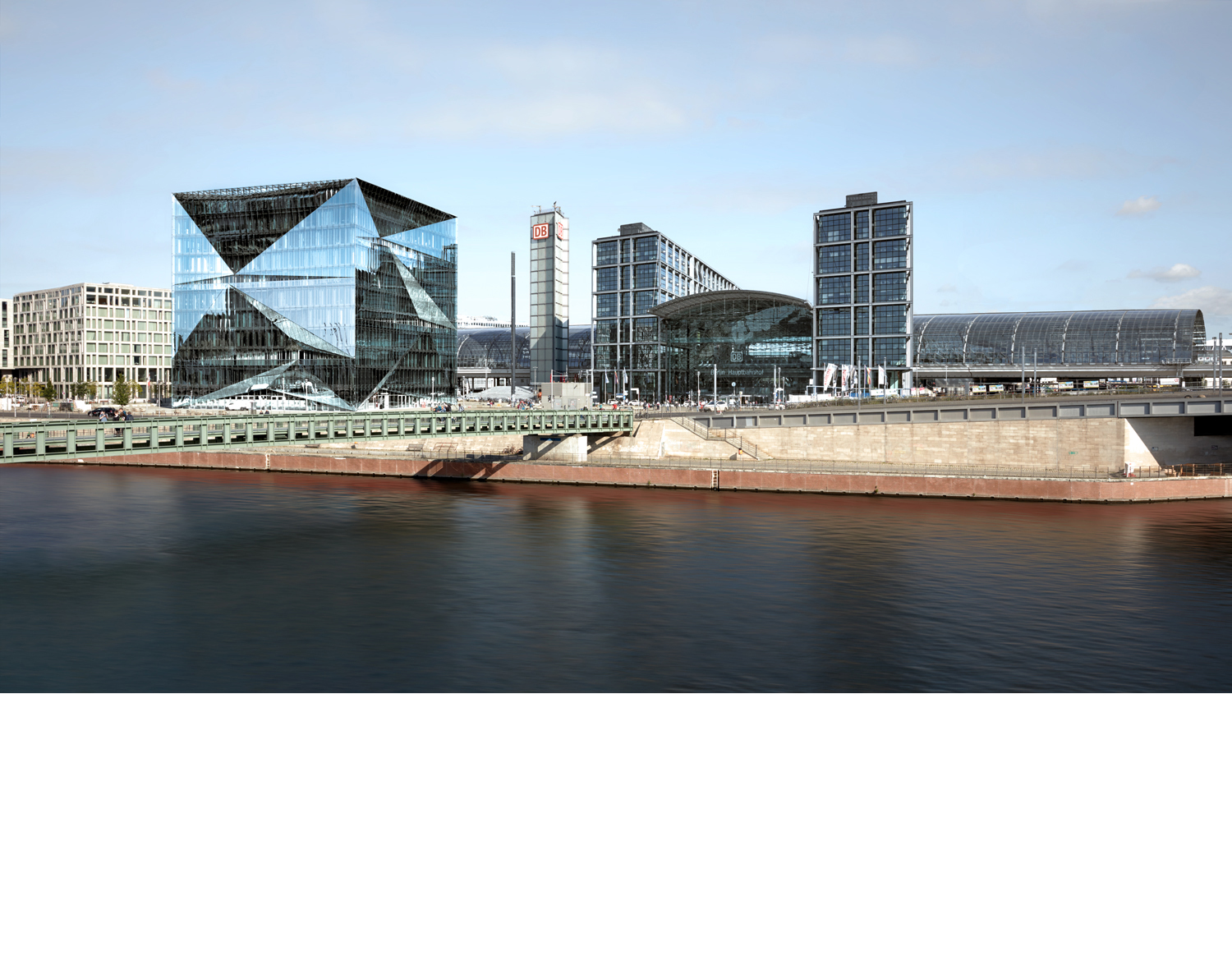 Aloys Kiefer Architekturfotografie: Querformat Aufnahme. Im Vordergrund ein Fluss. Links eine schmale Brücke, im Hintergrund links ein sehr großer Würfel mit gebrochener Glasfassade als Gebäude.